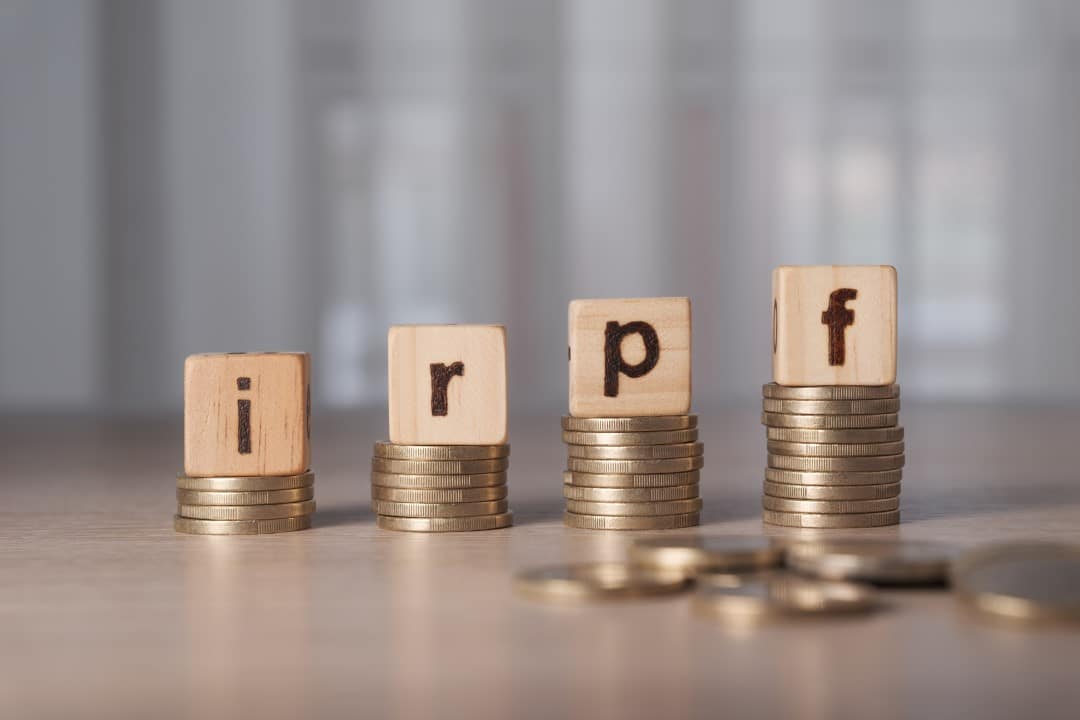 Blocos de madeira que soletram IRPF em pilhas de moedas, sigla Imposto de Renda Retido na Fonte