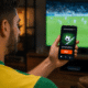 Homem em primeiro plano, visto de costas e levemente desfocado, segura um smartphone que exibe um aplicativo com a tela "Betting Online" e um botão laranja escrito "BET". Ao fundo, uma televisão exibe uma partida de futebol borrada. A imagem ilustra o cenário de apostas online, frequentemente associado a discussões sobre a regulamentação do mercado de apostas no país.